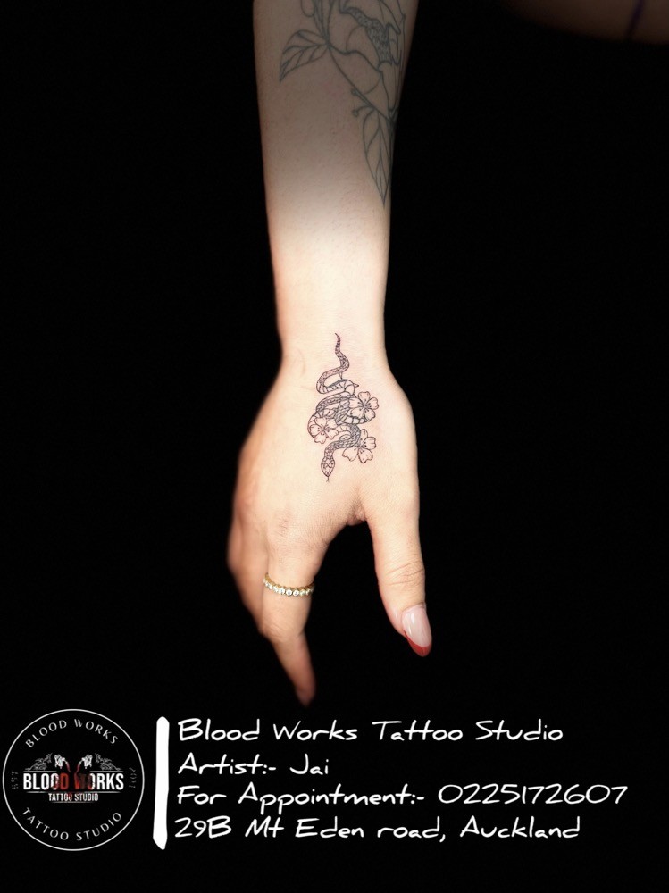 Finely detailed tattoo of a snake intertwined with delicate floral motifs on the wrist and hand area. Inked by Jai at Bloodworks Tattoo Studio, Auckland.