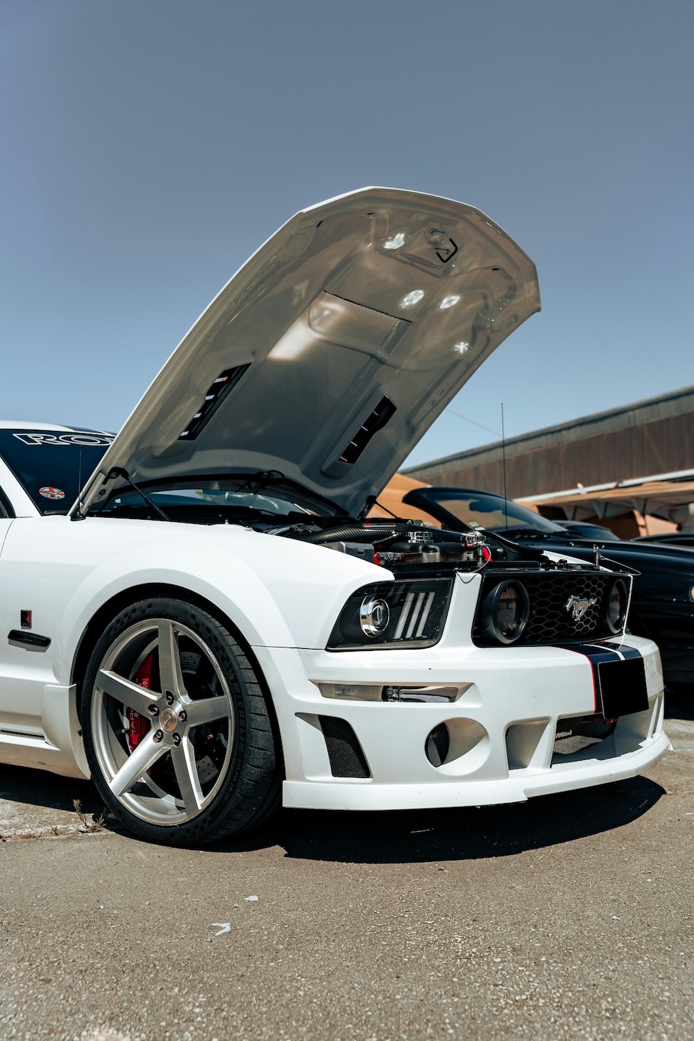 A sleek white sports car with its hood open is parked under a clear blue sky, showcasing its detailed engine components and shiny silver alloy wheels.