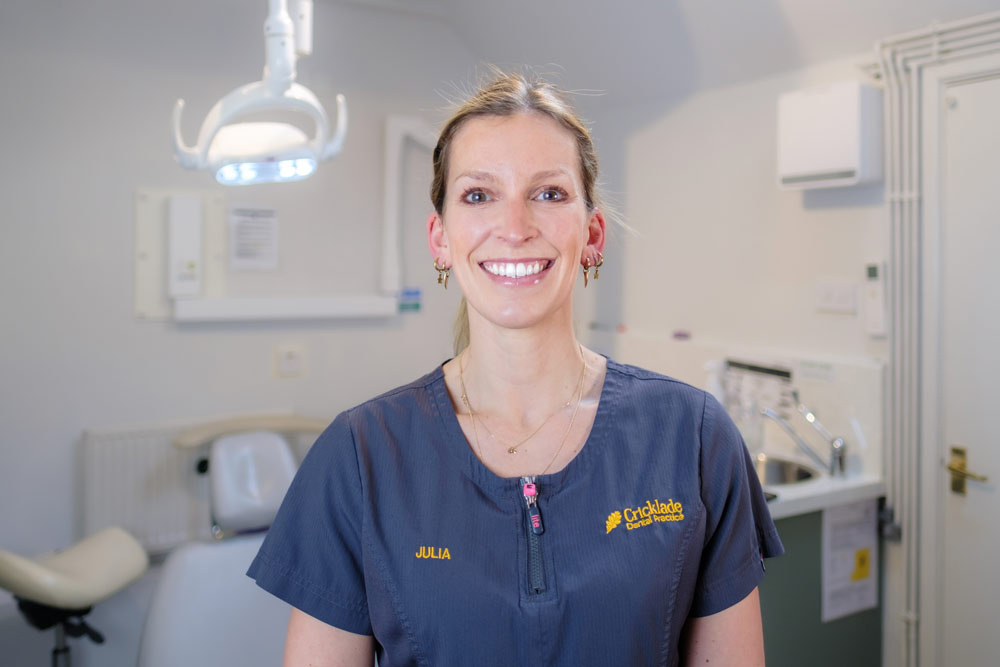 A portrait of Julia, Principal Dentist at Cricklade Dental Practice, smiling and wearing a dark grey scrub top with a pink zipper pull. The top features yellow embroidered text reading "JULIA" on the left. She is standing in a dental surgery with a dental light and white door visible in the background.