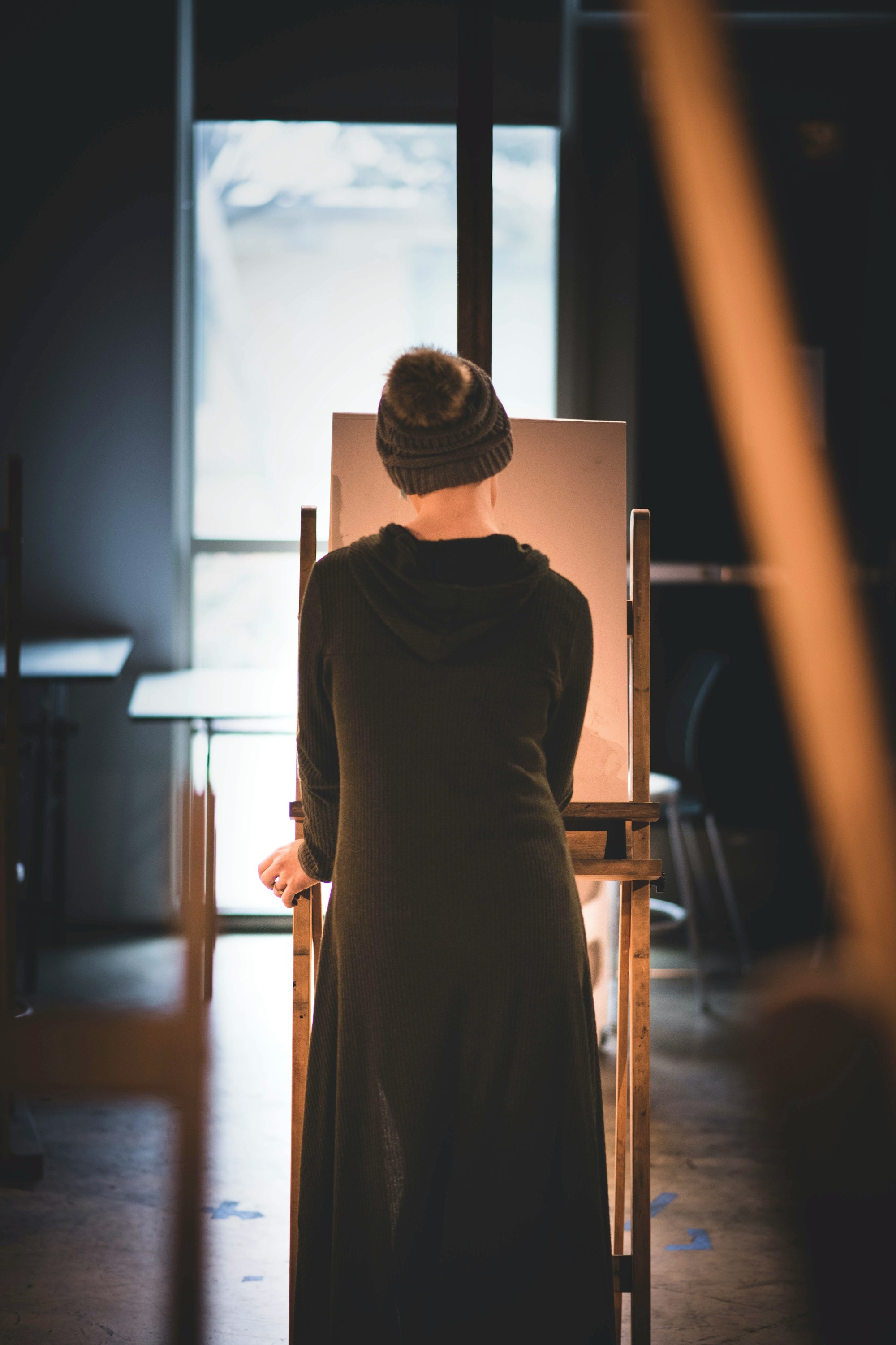Person seen from behind in a knit hat and dark sweater, working at an easel by a bright studio window