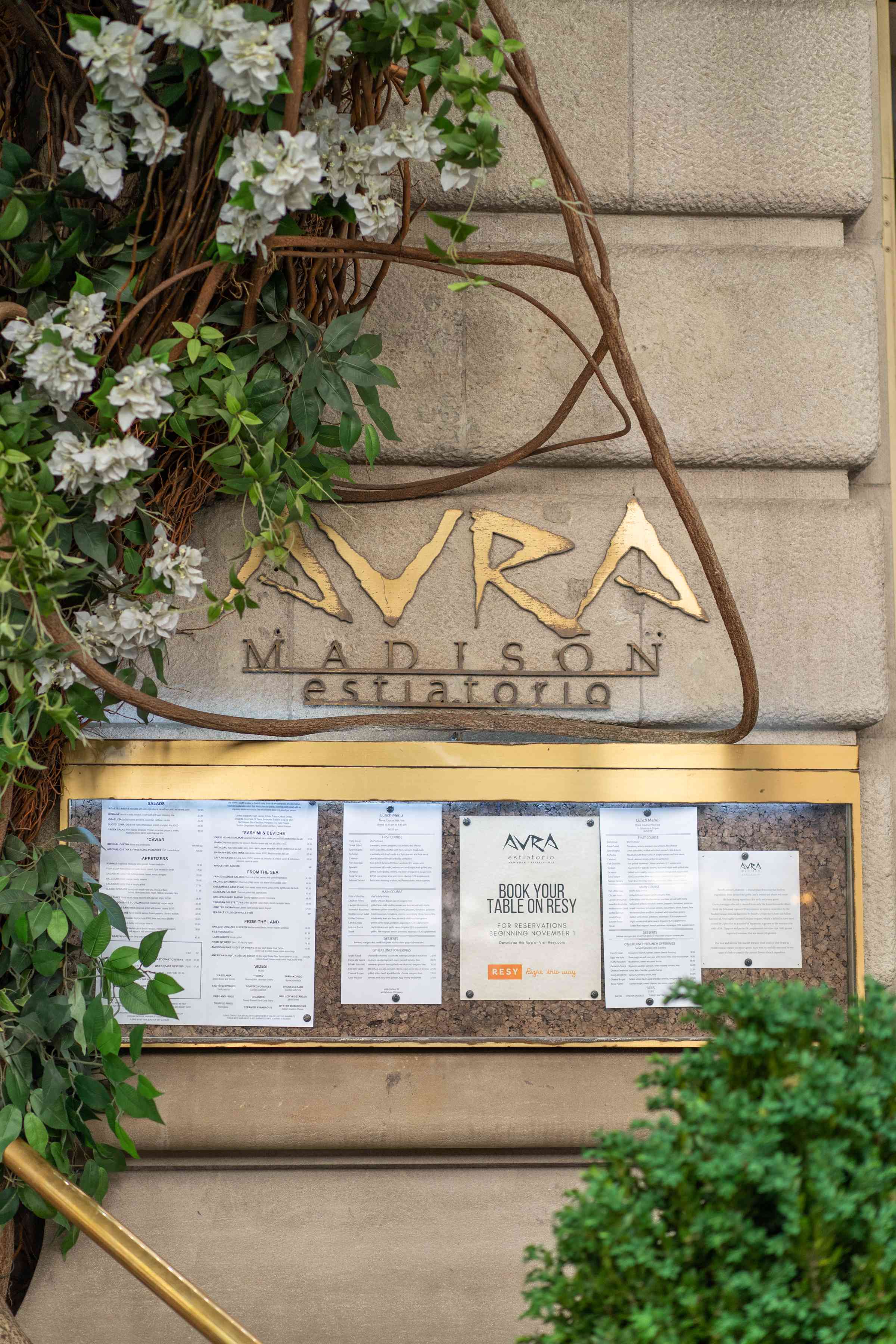 A decorative entrance featuring a sign and menus surrounded by green foliage and white flowers.