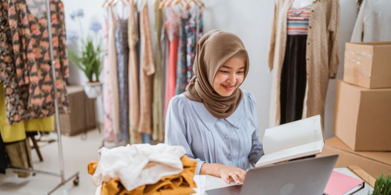 Peniaga butik wanita sedang uruskan stok inventori menggunakan laptop.