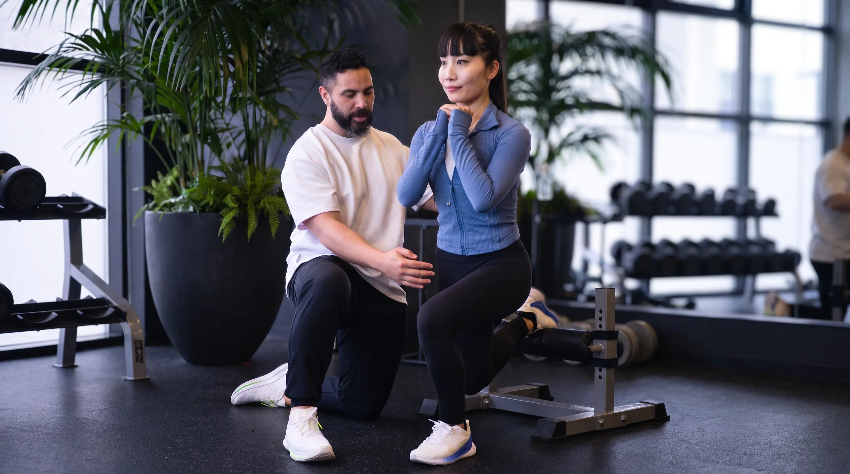 A coach kneels beside a woman at the bottom of a deadlift, guiding her head and back position before she lifts