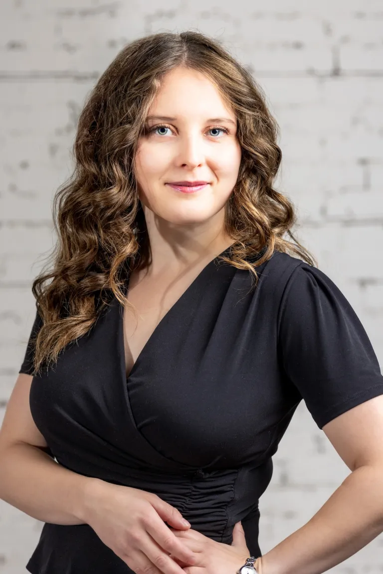 Image of Woman in black dress standing in front of white brick wall with a warm gentle smile.