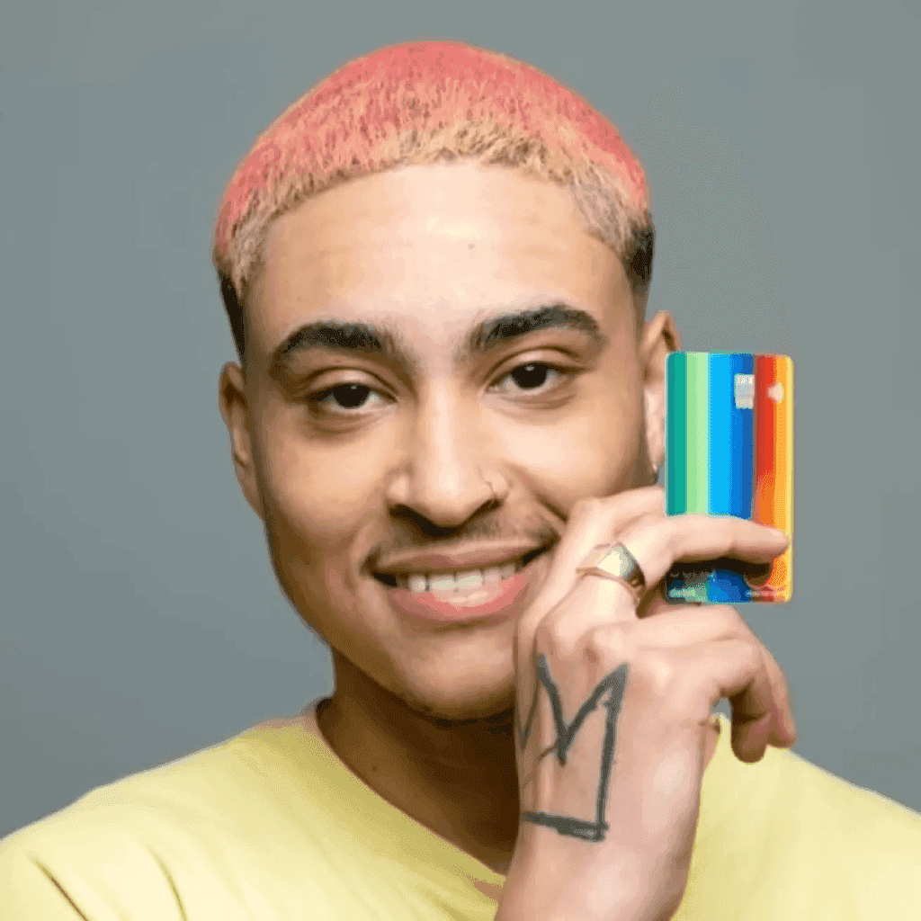 Een persoon met roze haar glimlacht terwijl die een kleurrijke kaart vasthoudt tegen een grijze achtergrond.