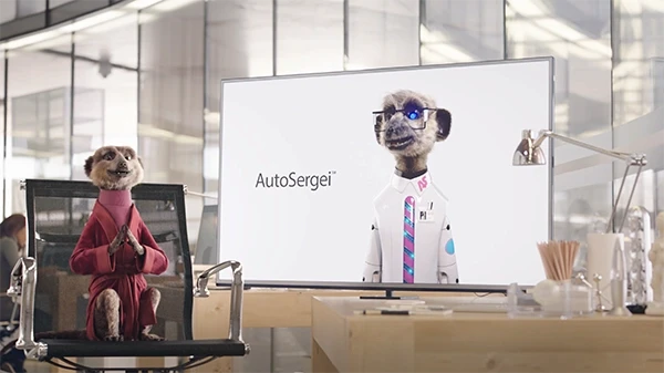 Compare the Market meerkat character in pink dressing gown watching AutoSergei AI meerkat on large office screen, Compare the Market TV advertisement still