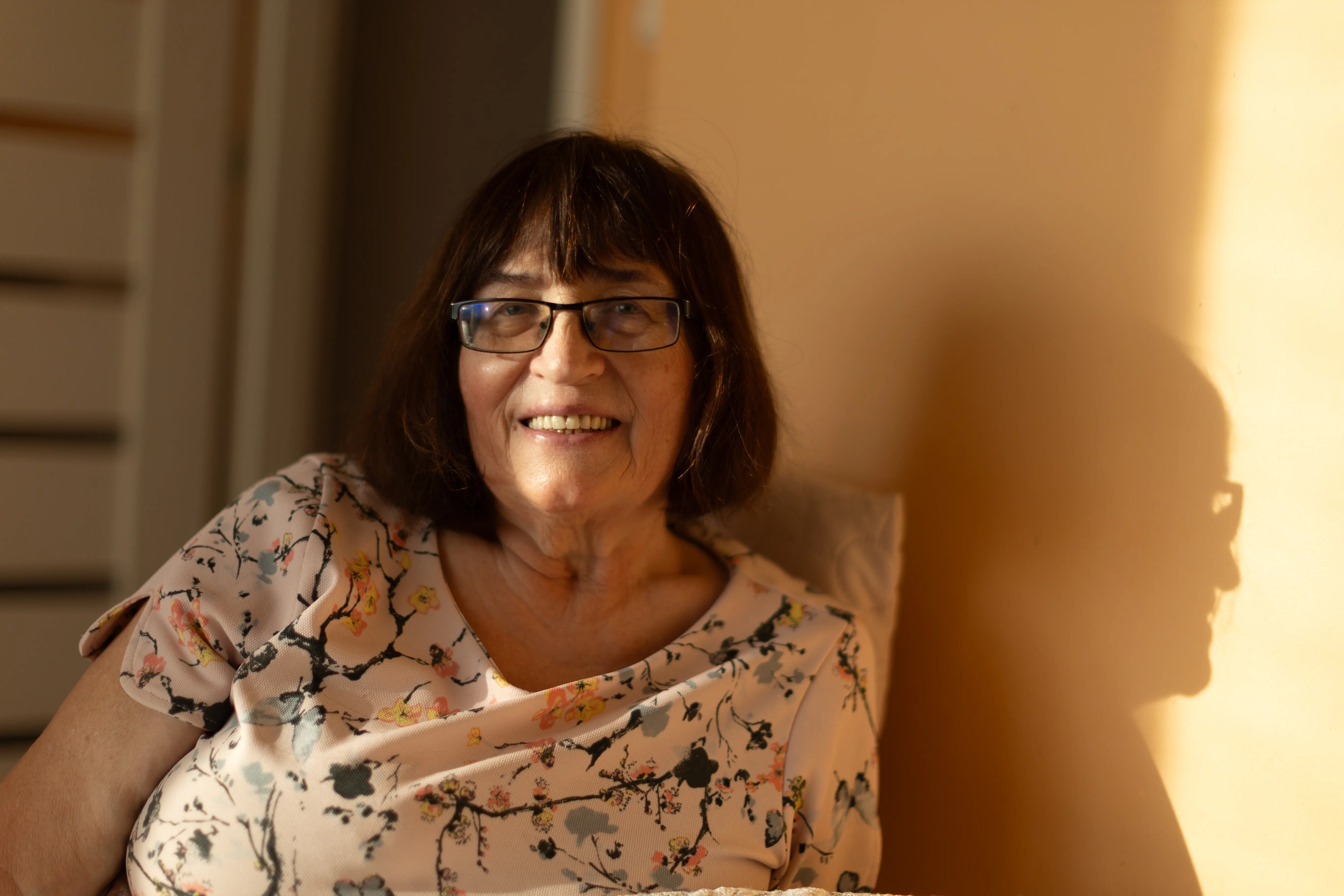 Portret uśmiechniętej kobiety o bąbkowych włosach i grzywce w okularach. Jest ubrana w jasną bluzkę. Padają na jej twarz promienie słońca.