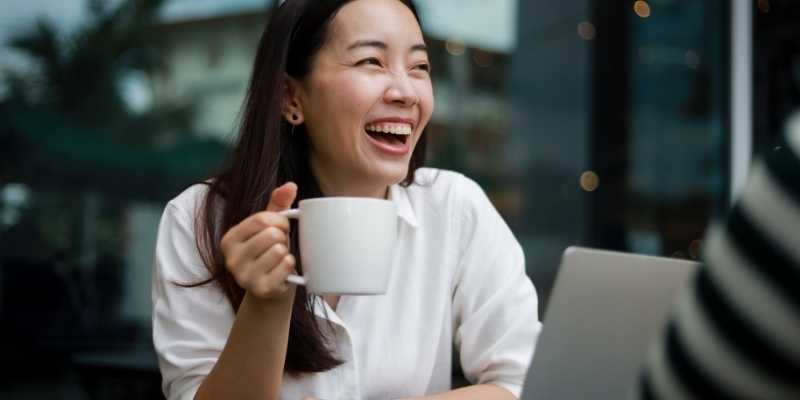 Asian girl laughing while holding a coffee cup in hand in a cafe.