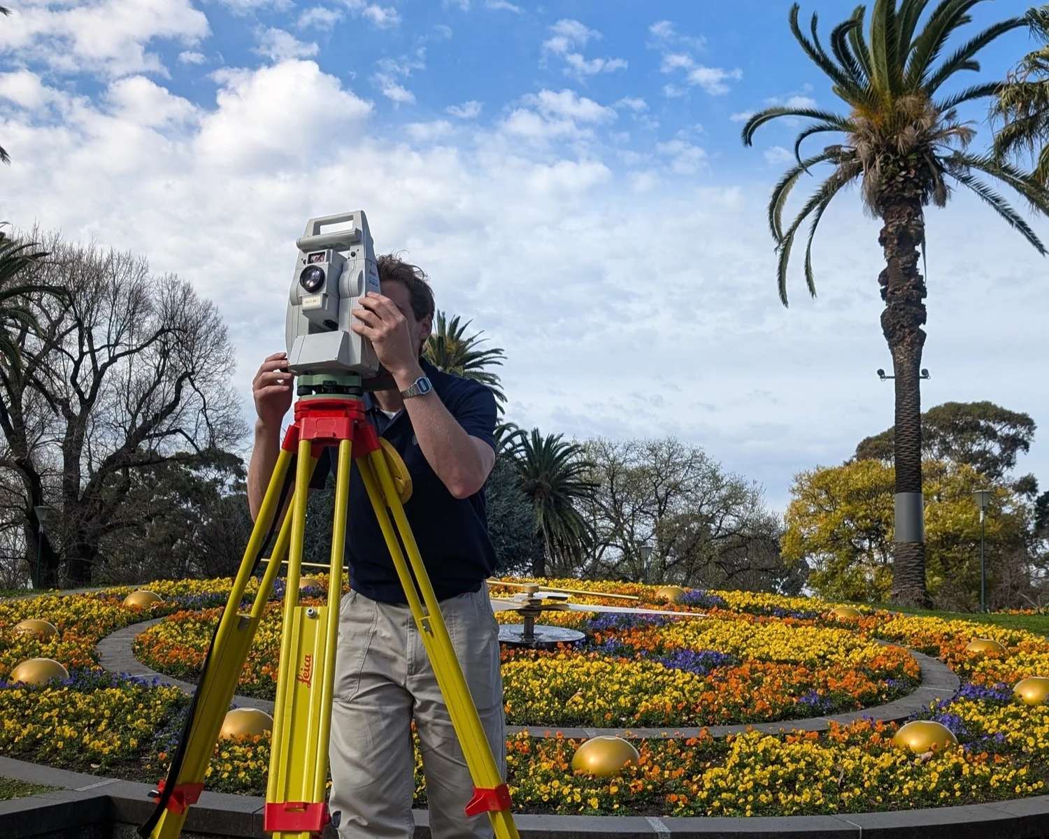 Land surveyor using a total station on a tripod in a landscaped public garden with palm trees and flower beds.