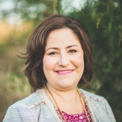A smiling woman with medium-length brown hair wearing a light jacket and a pink top, standing outdoors.