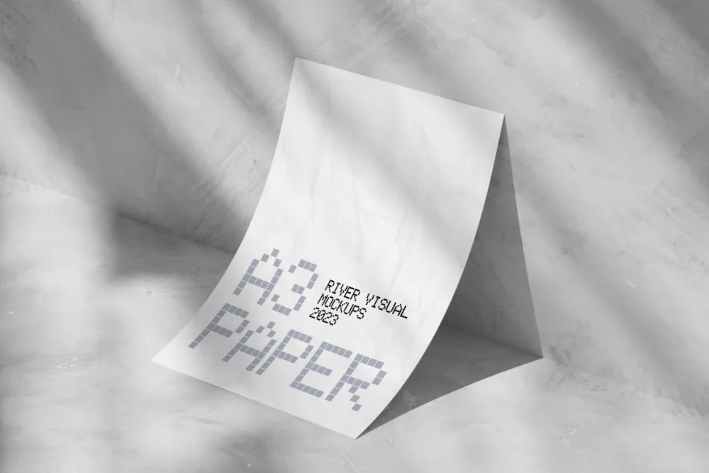 An A3-sized paper mockup with a crumpled texture, displayed in a minimalist setting. The paper is bent and placed on a concrete-like surface, illuminated by soft natural light with subtle shadows.