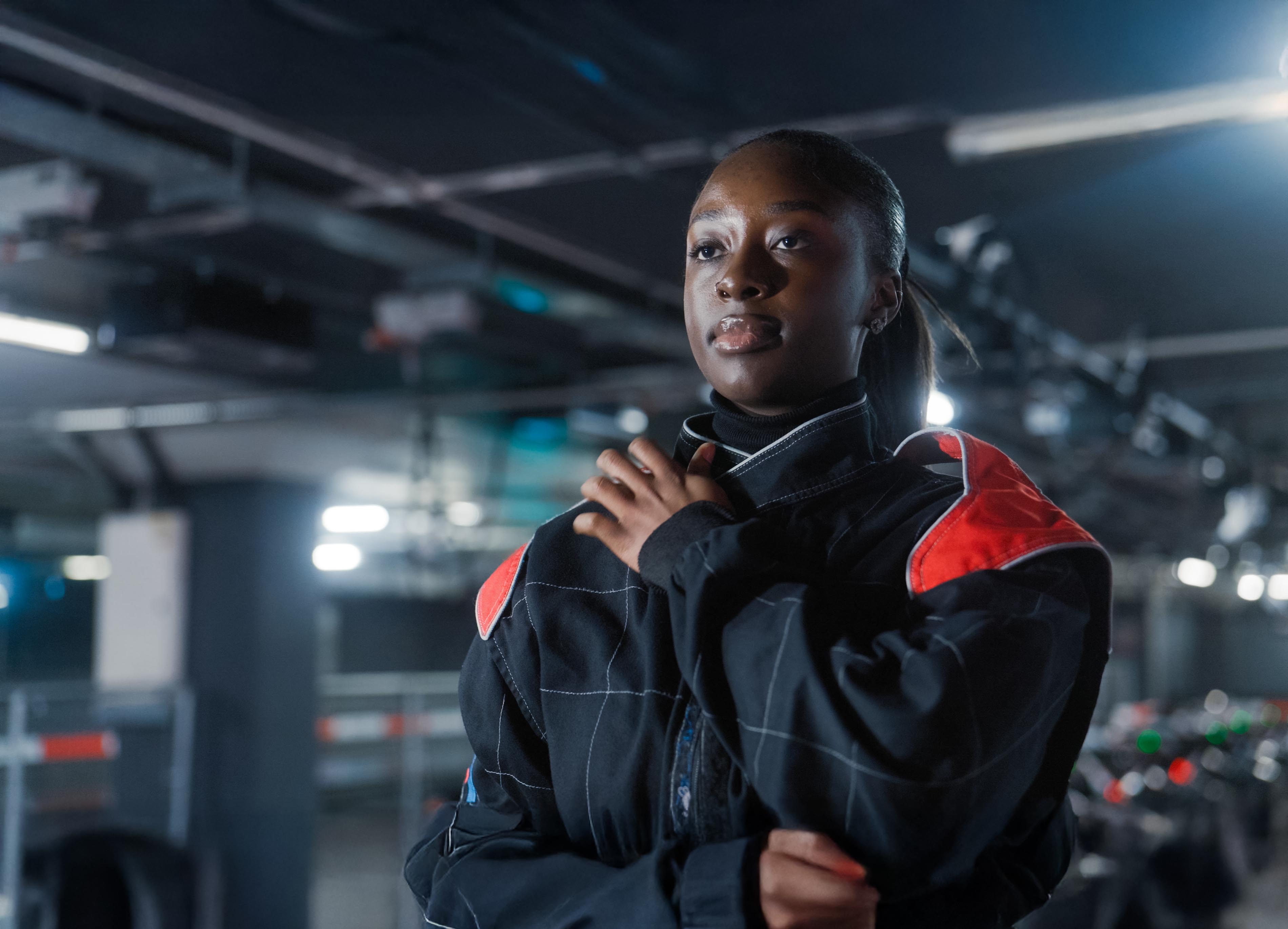 A young Black woman in a black racing suit looks thoughtfully to her right in a dimly lit garage.