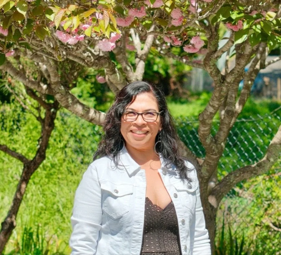 Uma mulher de óculos e cabelo longo e escuro está sorrindo sob uma cerejeira em plena floração, vestindo uma jaqueta branca e uma blusa de renda preta, com um exuberante jardim verde ao fundo.