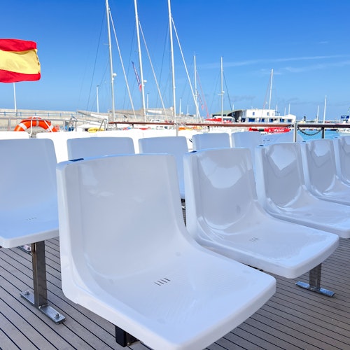 White plastic seats on a boat deck with a Spanish flag and several docked sailboats in the background under a clear blue sky.