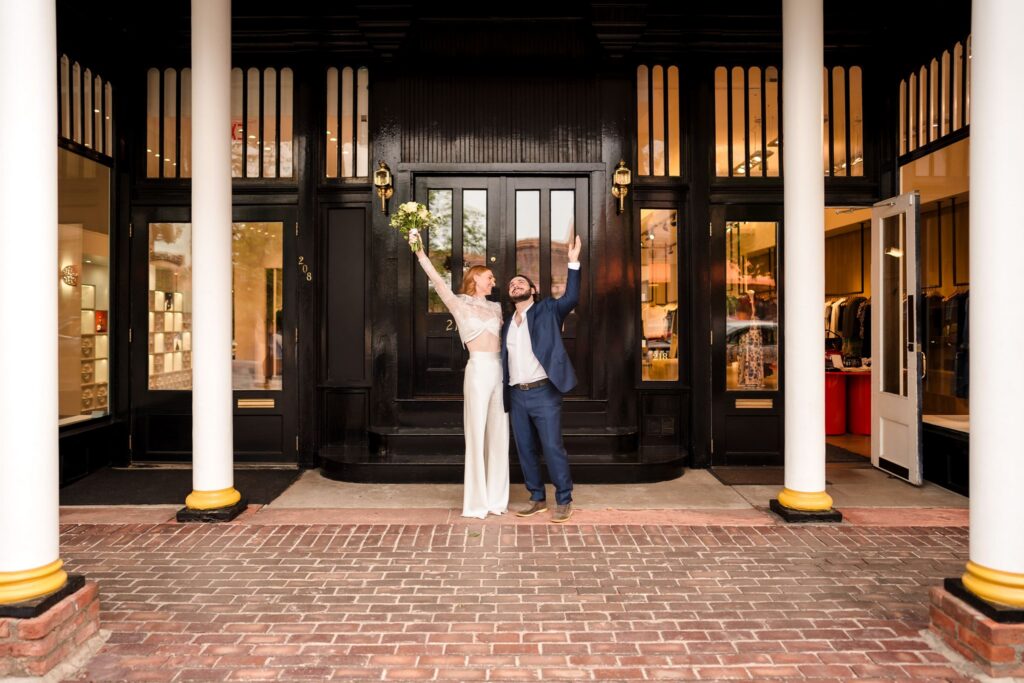 Wedding couple raising their arms outside a dark doorway framed by tall white columns