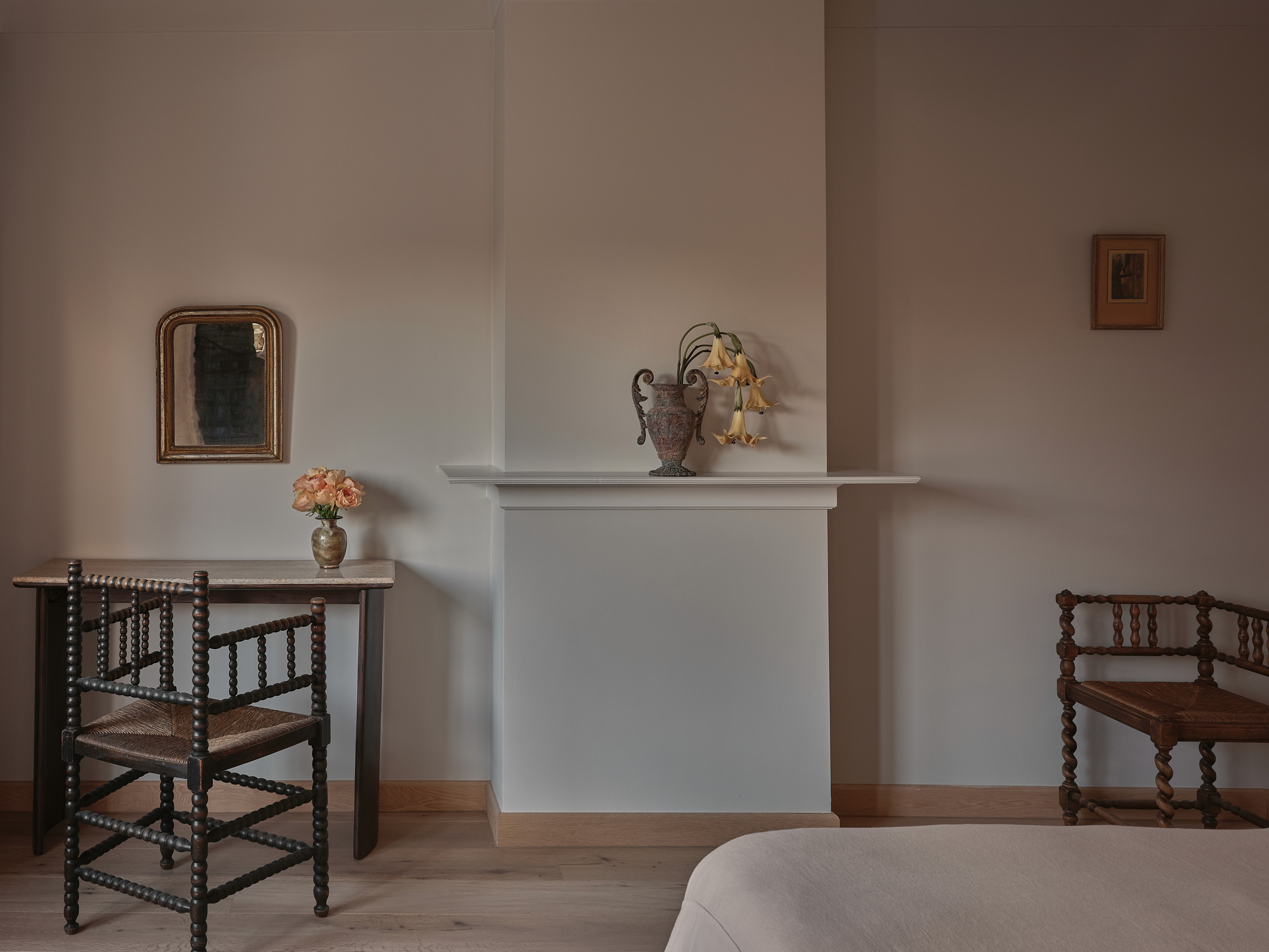 Bedroom with antique bobbin chairs, white fireplace mantel with decorative urn and dried flowers, gilt-framed mirror, and fresh roses on console