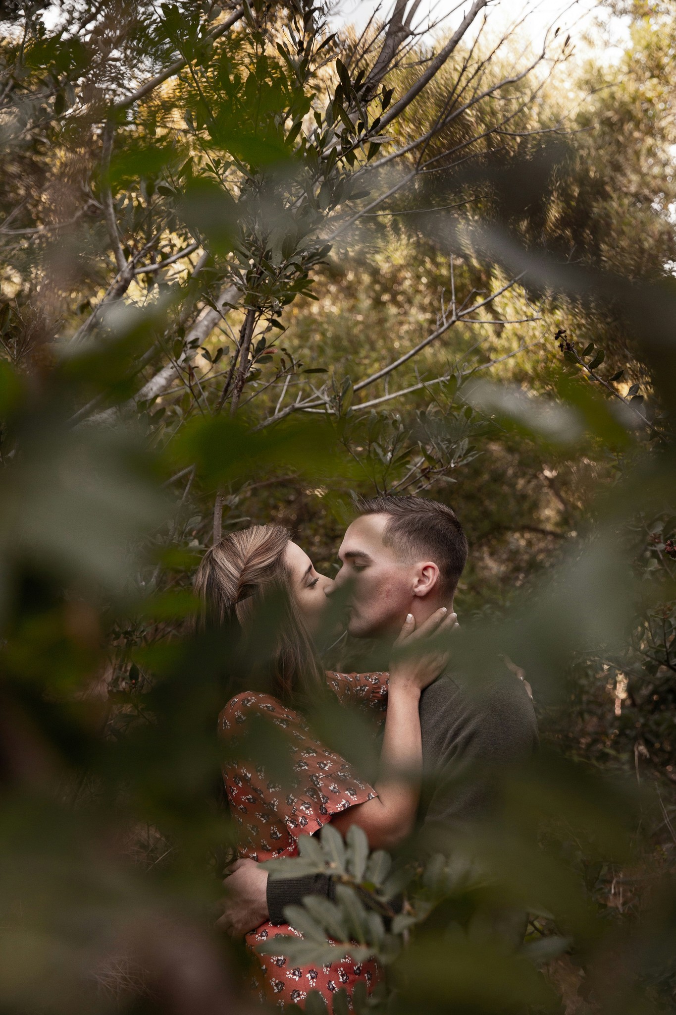 Couple sharing a kiss framed through lush green foliage during an intimate outdoor engagement session