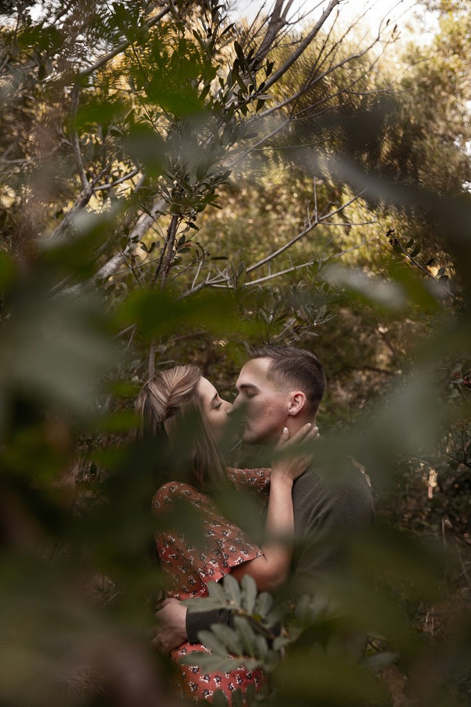 Intimate moment of a couple kissing among dense green foliage with soft golden light filtering through the trees