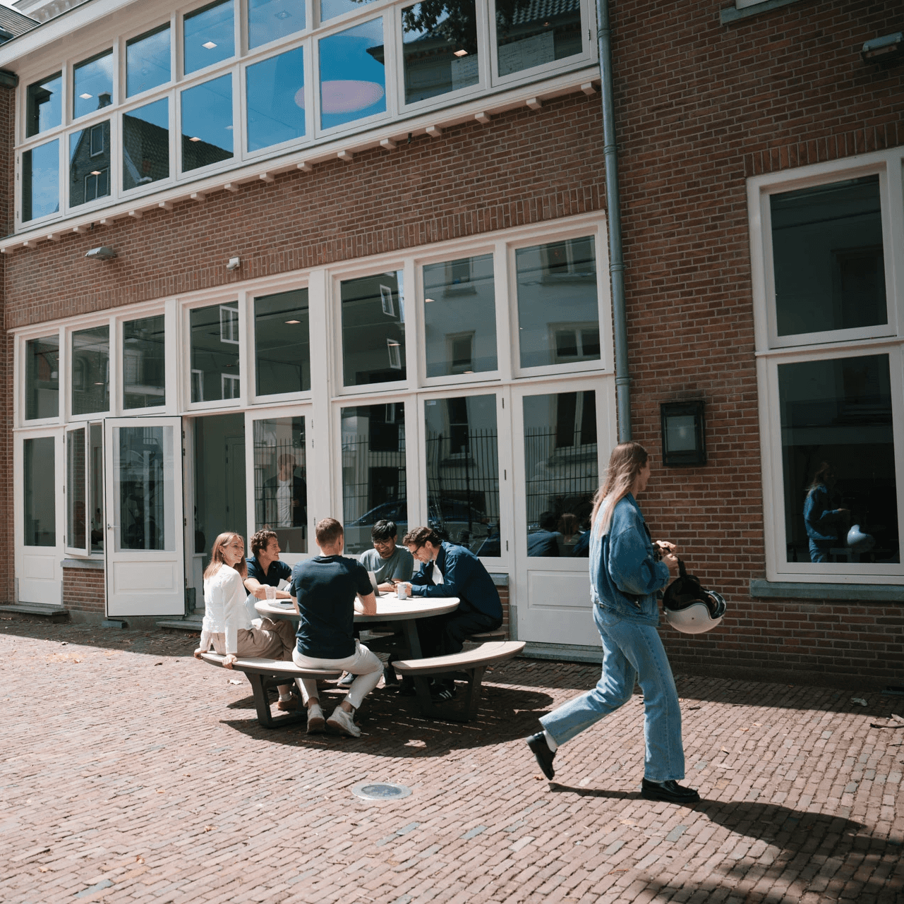 A group of people sits around a table outside a building on a sunny day, while another person walks by.
