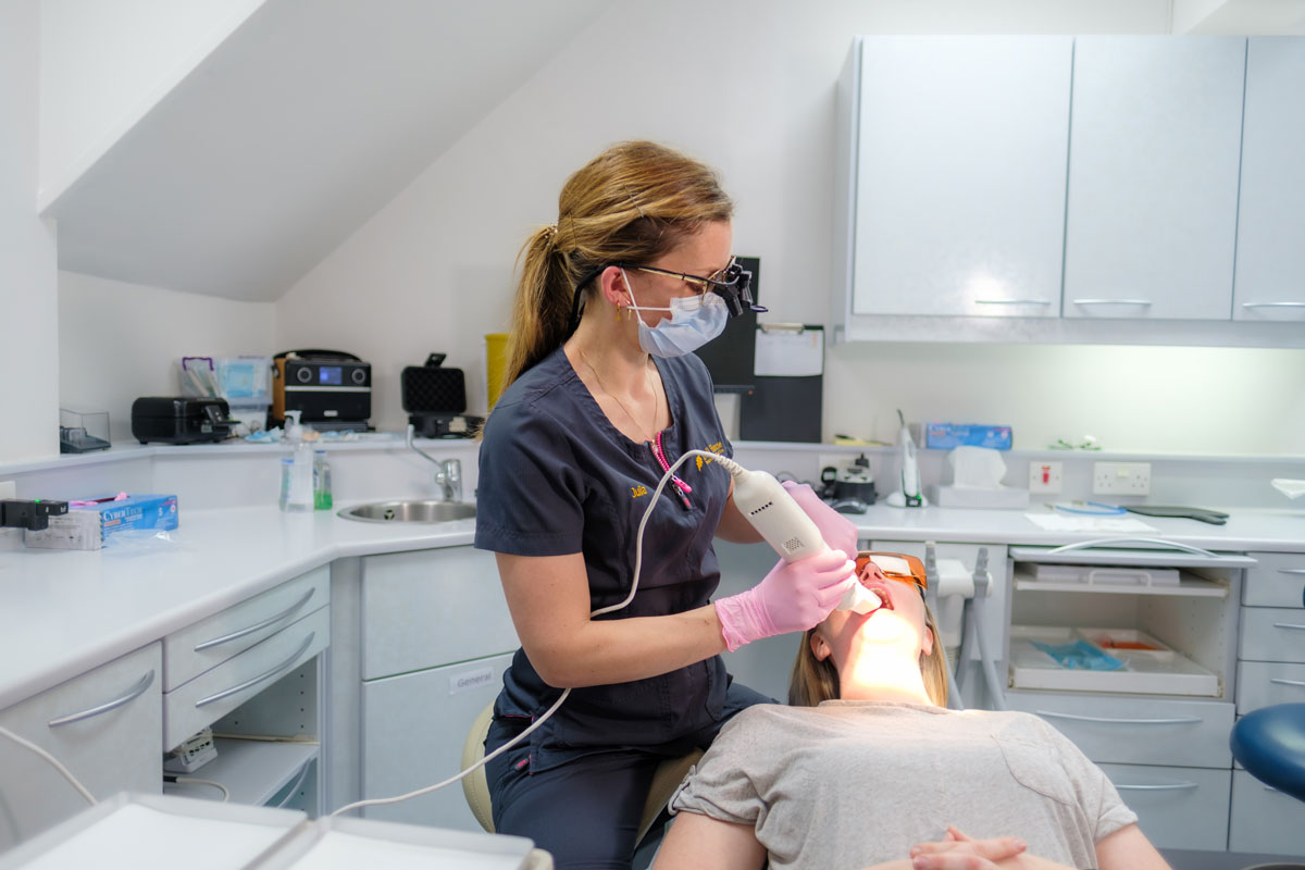 A dentist wearing a face shield and mask uses an intraoral scanner on a patient to create a digital impression. The patient, also wearing protective eyewear and a cheek retractor, lies in a dental chair.