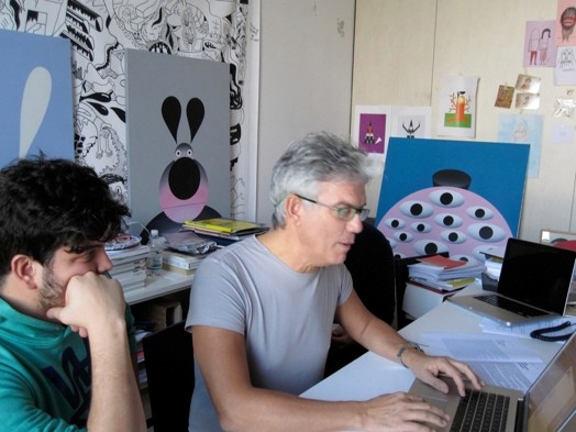 A photo of two people working at a laptop in an art workshop filled with canvases of drawn cartoons.