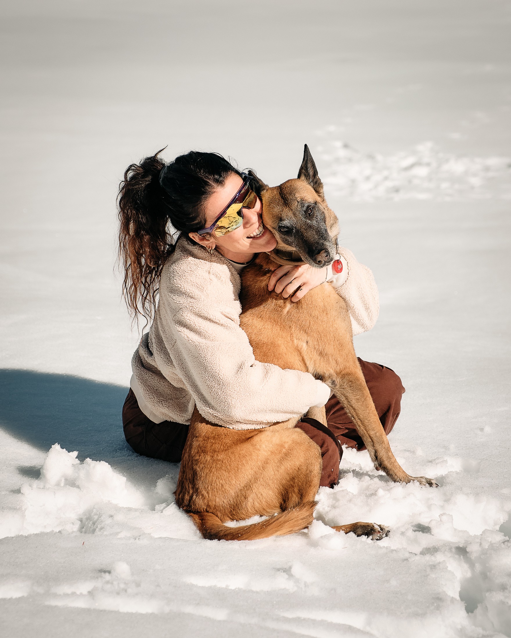 A woman in sunglasses happily hugs her dog in a snowy landscape, showcasing a joyful bond between them.