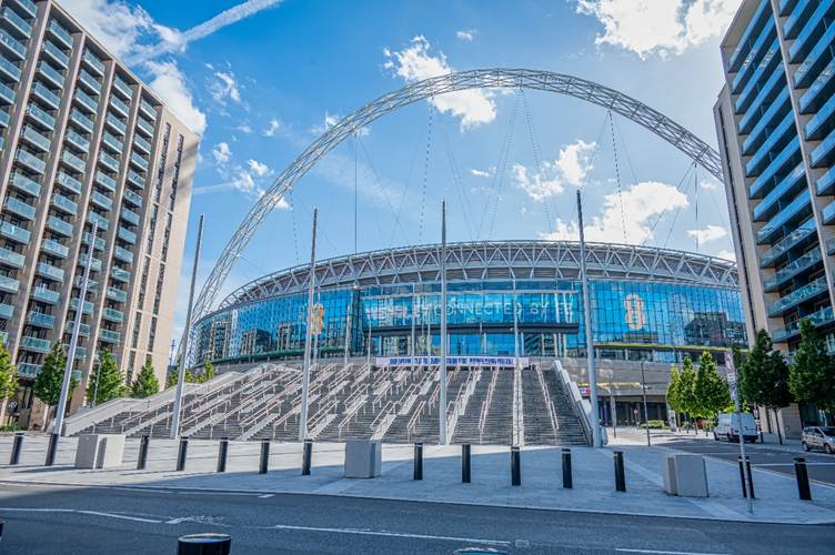 The image shows a modern stadium with a distinctive roof structure, surrounded by contemporary buildings and an empty street, under a clear blue sky.

AI-generated content may be incorrect.