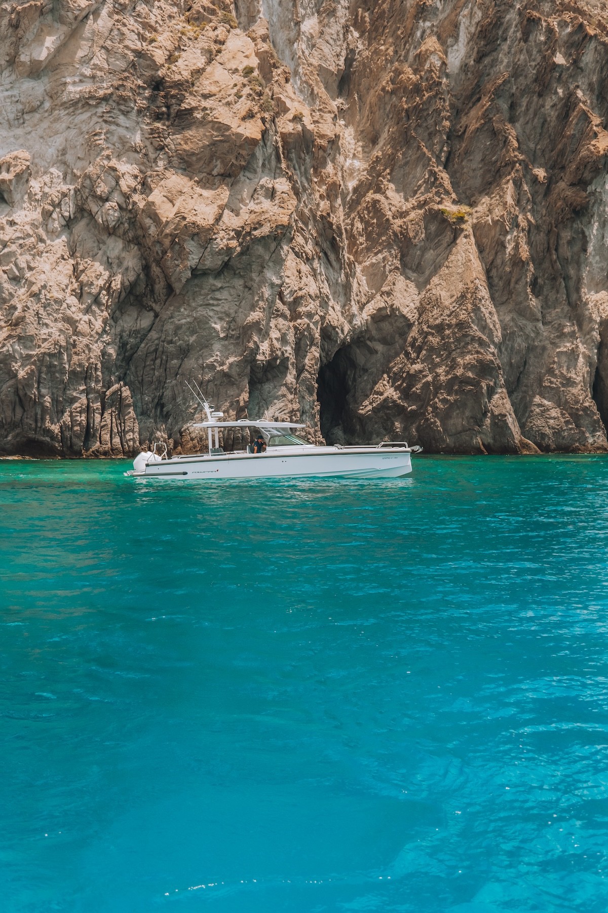 White Axopar 37 yacht Anima anchored in crystal-clear turquoise waters against dramatic white limestone cliffs in the Cyclades.