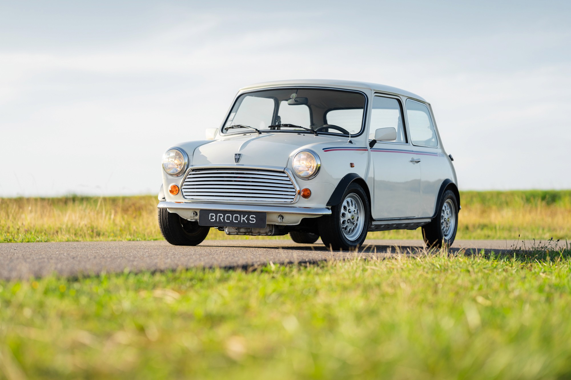 A vintage green mini classic automatic car drives along a road, surrounded by fields and a clear blue sky in the background.