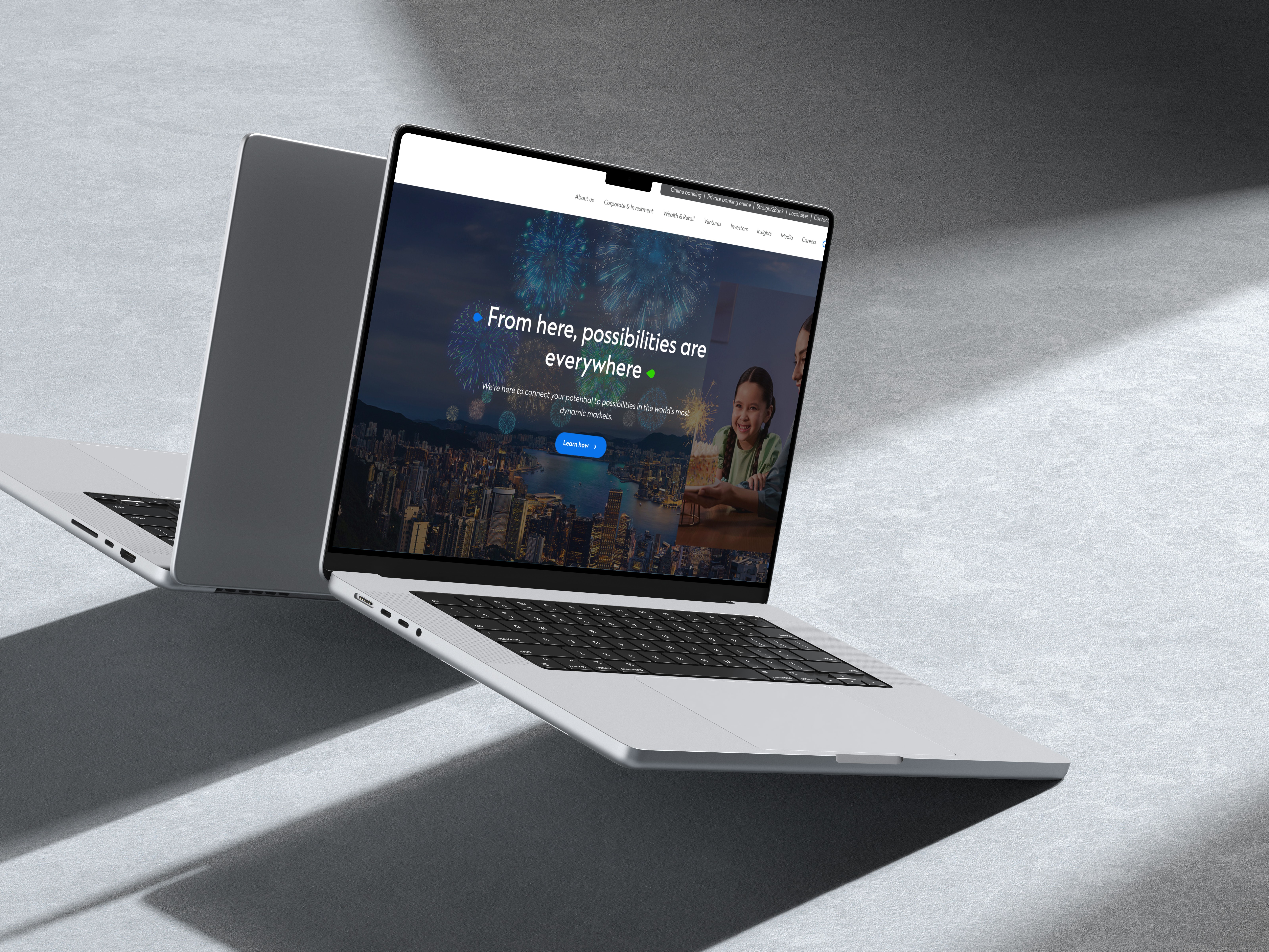 Macbook Pro sur une surface grise affichant la page d'accueil du site bancaire. L'écran montre une vue nocturne de la ville avec des feux d'artifice et un menu de navigation. Un simple ordinateur portable blanc placé en biais crée des ombres nettes.