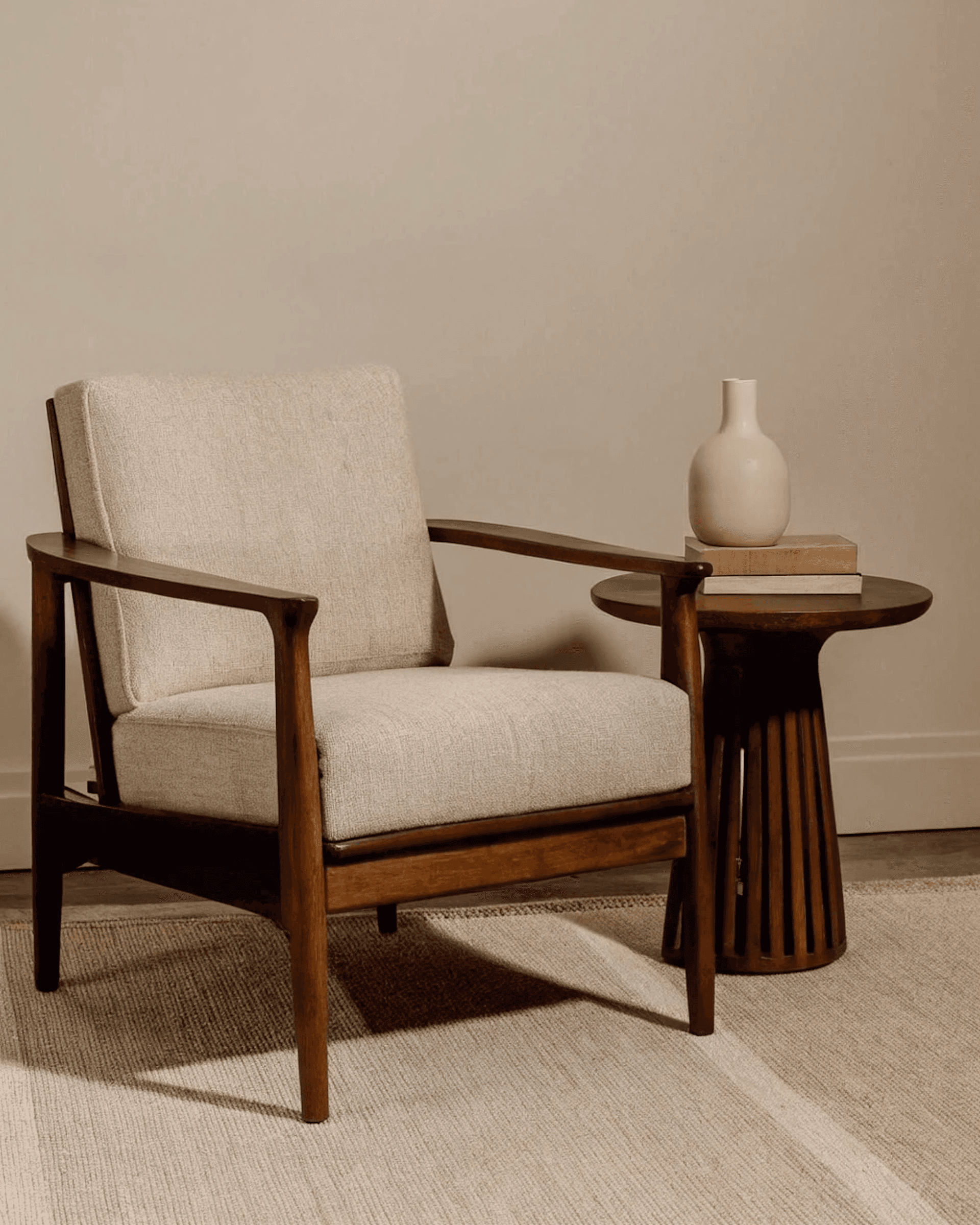 Mid-century modern armchair with beige cushions, paired with a wooden side table holding a vase and books, set on a woven rug.