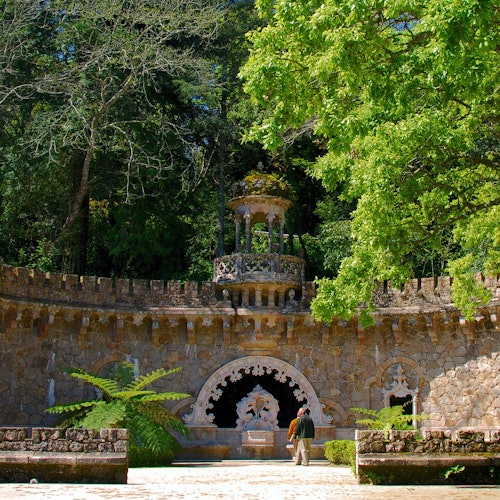 一个人在花园环境中行走,背景是一堵有拱形开口的石墙,以及装饰性的结构和郁郁葱葱的绿植。