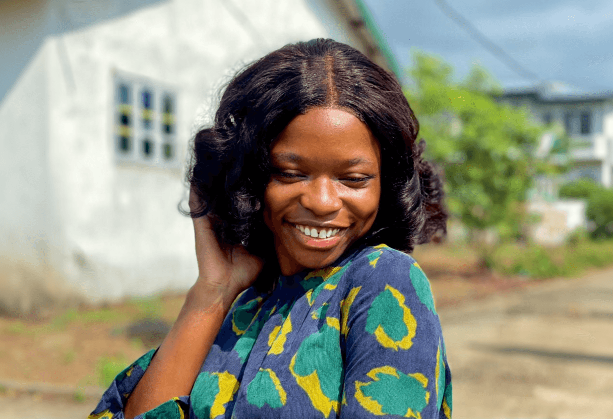 Outdoor portrait of a smiling young woman with shoulder-length wavy black hair, looking down while touching her hair, wearing a blue dress with green and yellow patterns, with houses and trees blurred in the background.