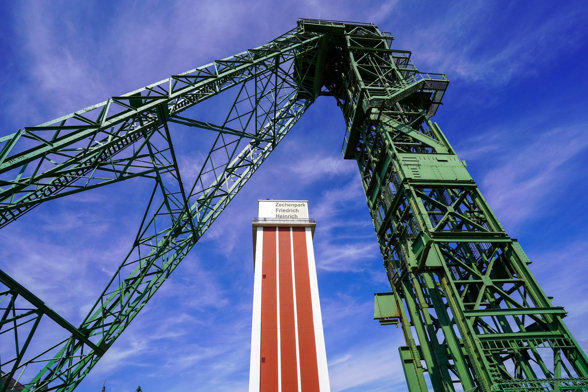 Perspektive von unten auf den roten Zechenturm. Der Turm steht zwischen den stahlroten Stützbeinen der Förderanlage. Über ihm ein blauer Himmel mit leichten Schleierwolken