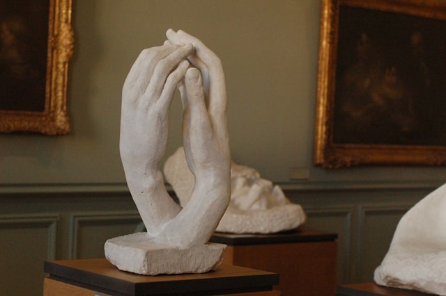 White marble sculpture of two hands delicately touching, displayed in a softly lit museum room adorned with classical paintings.