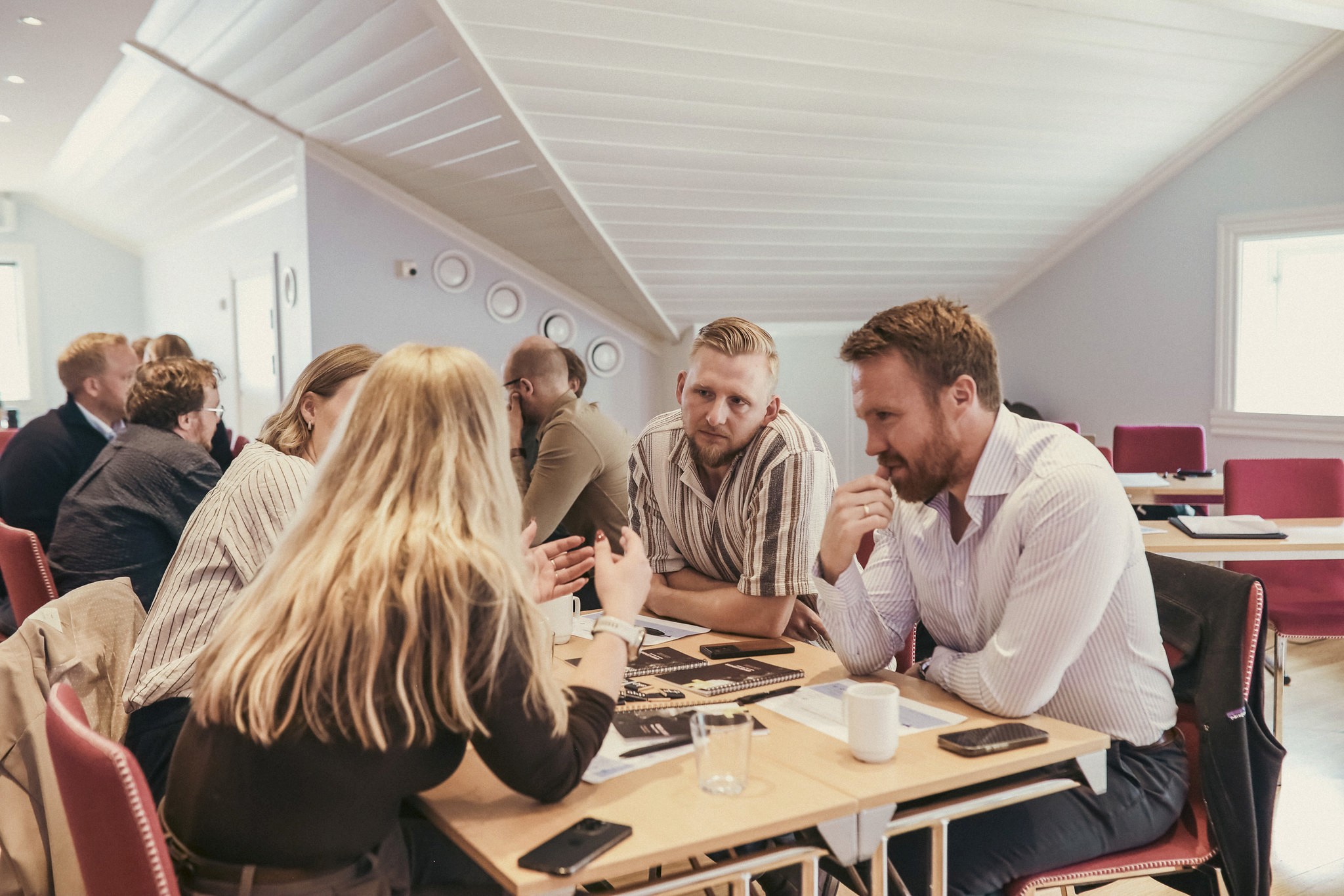 A woman with blonde hair smiles in a social setting, with people blurred in the background. Bright window visible.