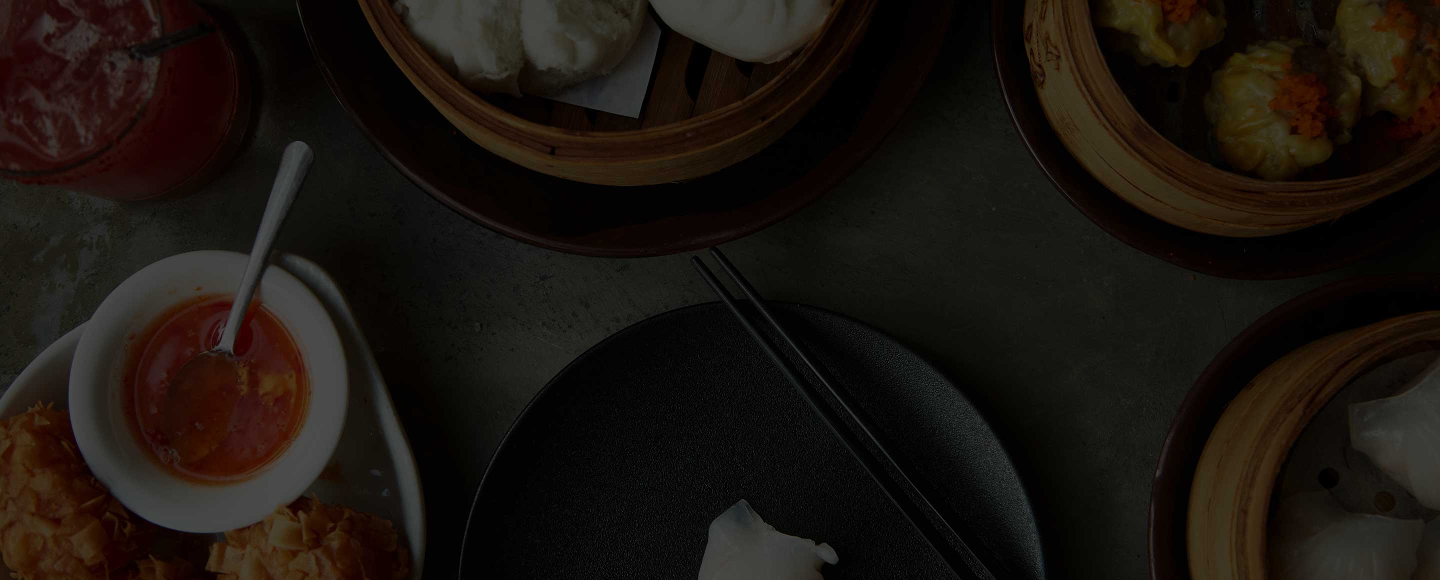 Top-down view of a dim sum spread featuring bamboo steamers, dipping sauce, and black chopsticks on a dark table setting.