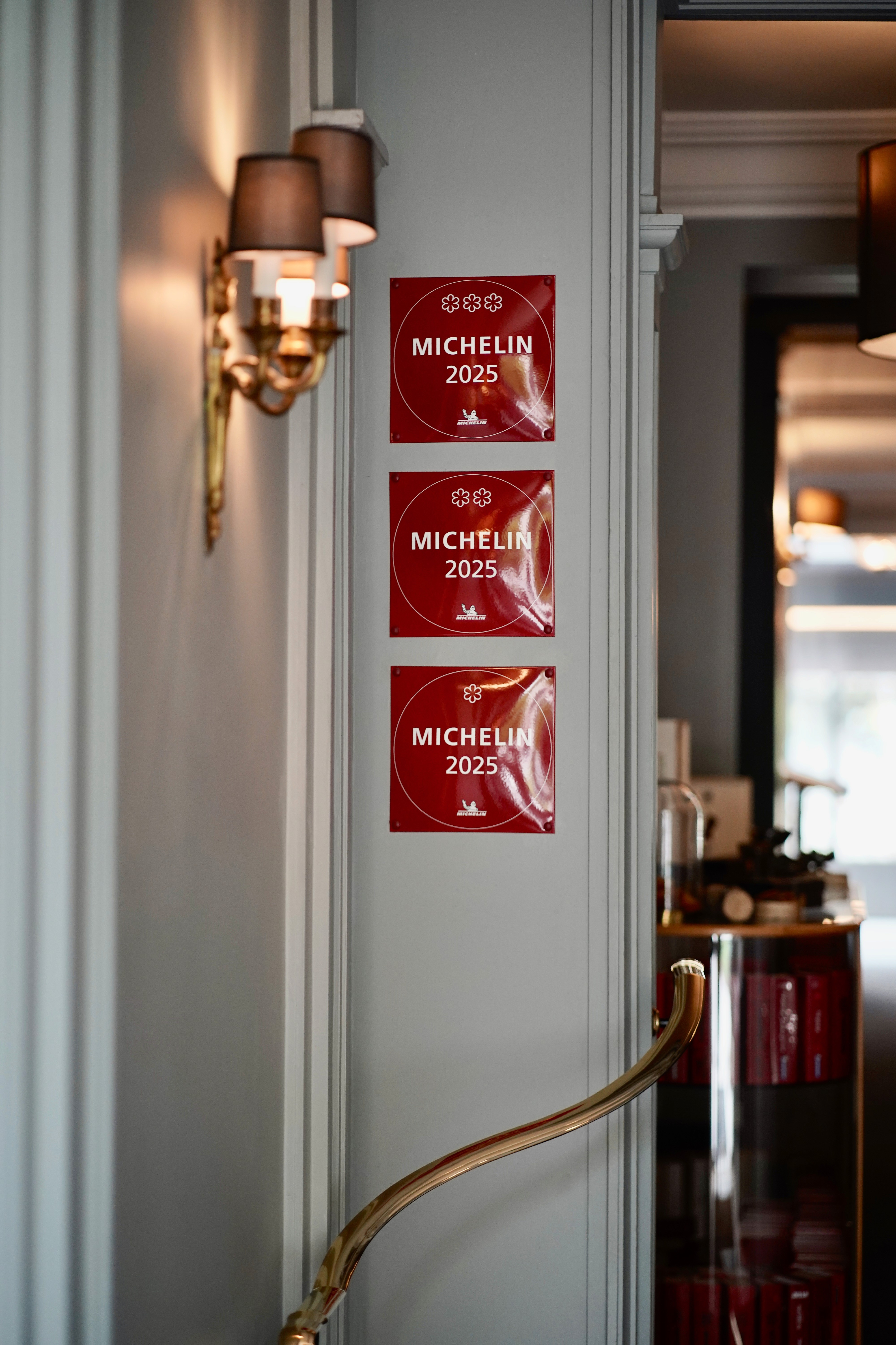 Vue détaillée de trois plaques Michelin millésimées 2025, affichées verticalement sur un mur gris dans l'entrée chic d'un établissement, montrant successivement une, deux et trois étoiles.