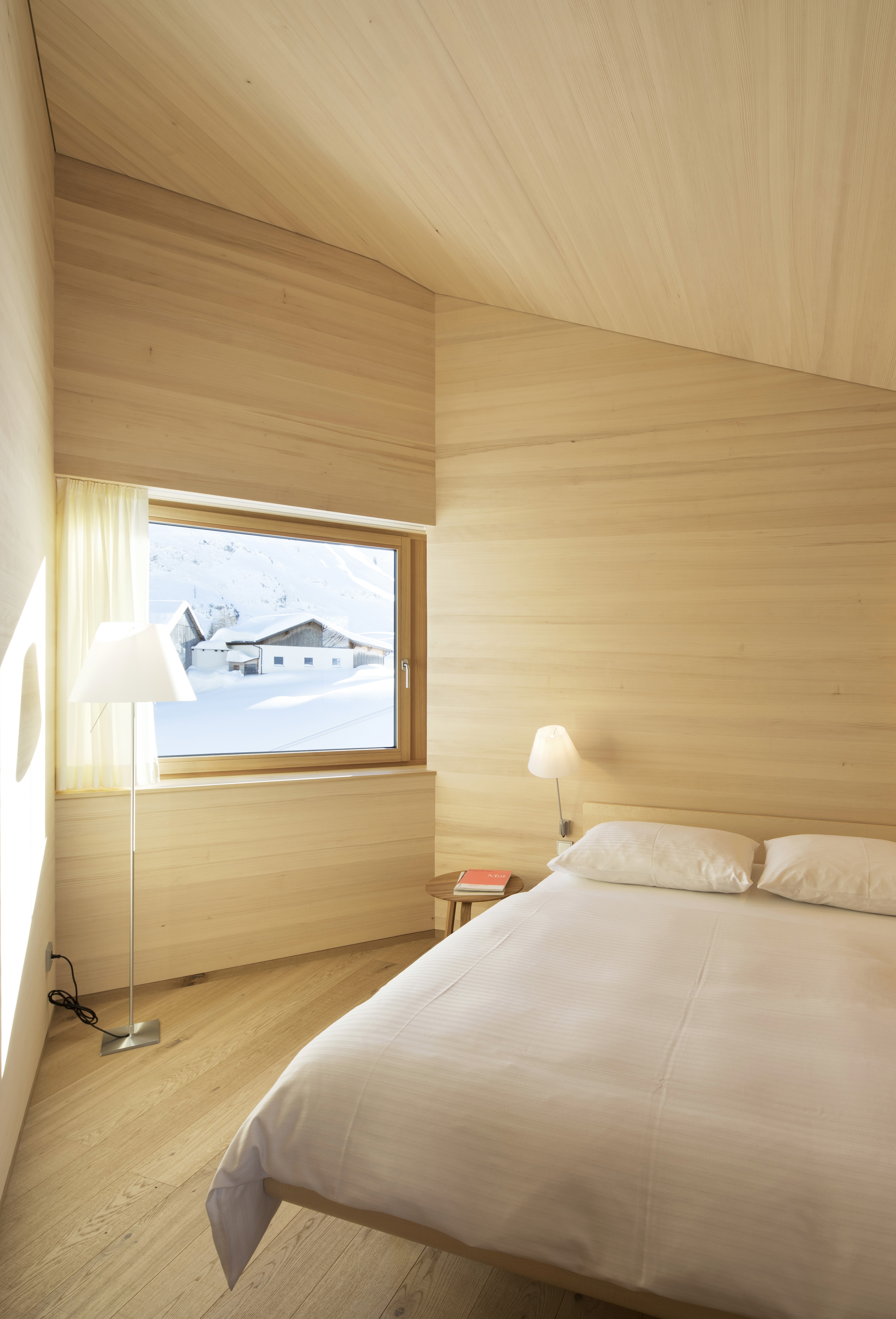 A bed in a wooden bedroom with a window looking out into the snowy landscape in Warth.