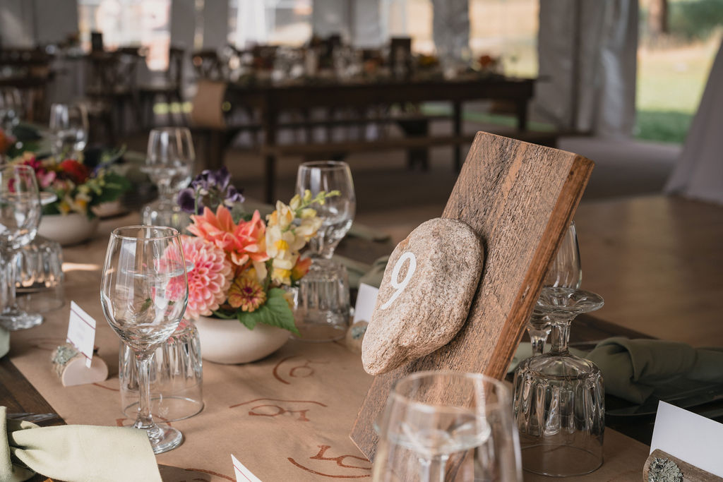 A close-up of a wedding reception table setting featuring a rustic wooden table number "9" painted on a stone. Soft pink and yellow flowers in a white vase decorate the table.