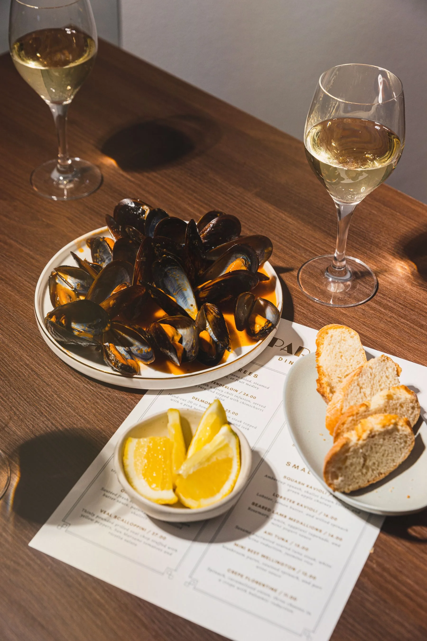 an overhead shot of a table with two glass of wine, a plate of mussels and bread, lemons, and a menu