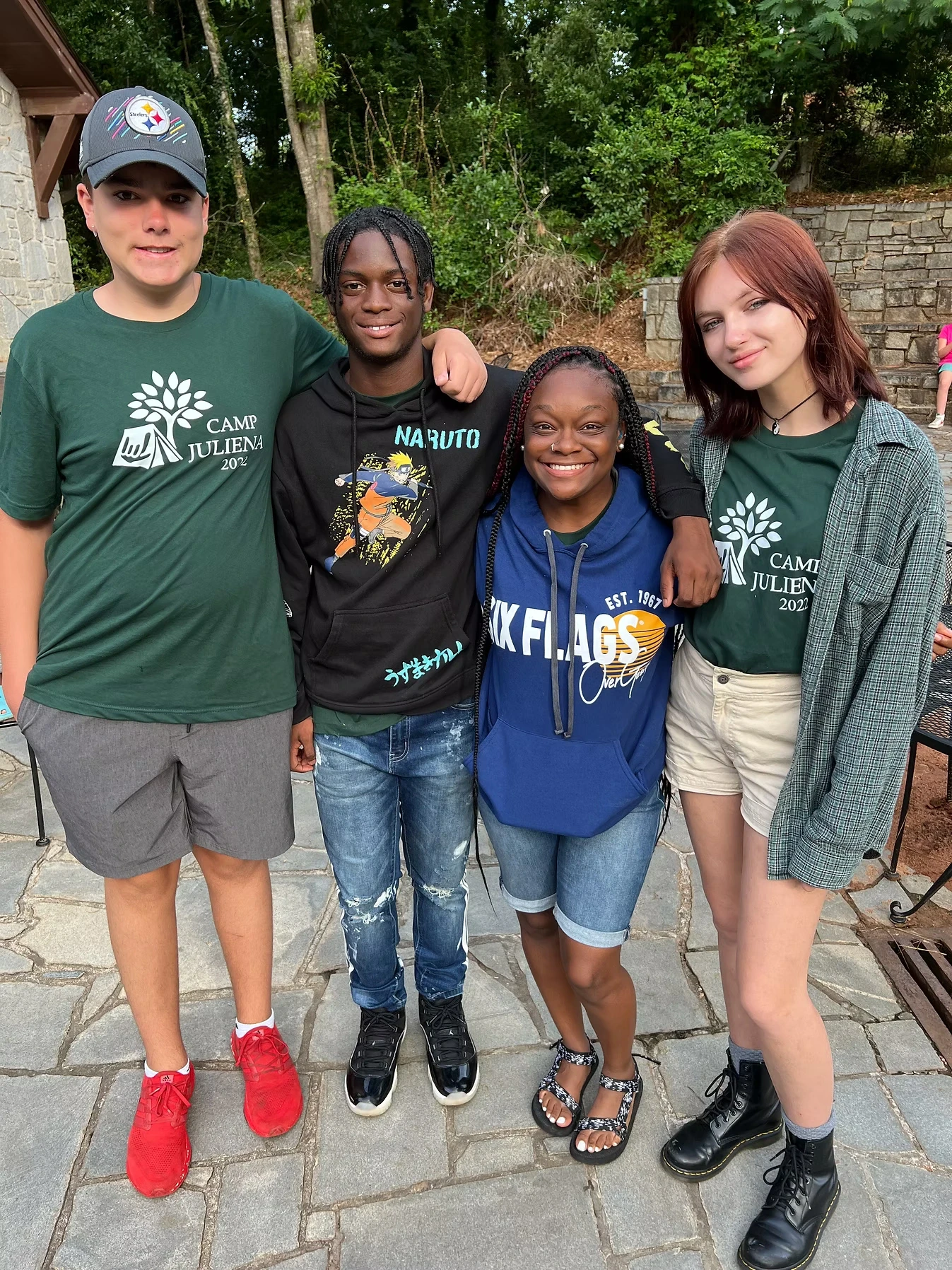 Four teenagers, two boys and two girls, stand together smiling for a photo. They're wearing casual clothes and two are wearing Camp Juliena t-shirts.