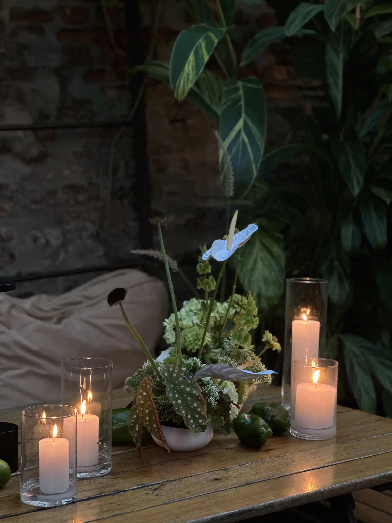 Centro floral con velas encendidas y follaje tropical sobre mesa de madera en ambiente íntimo y cálido.