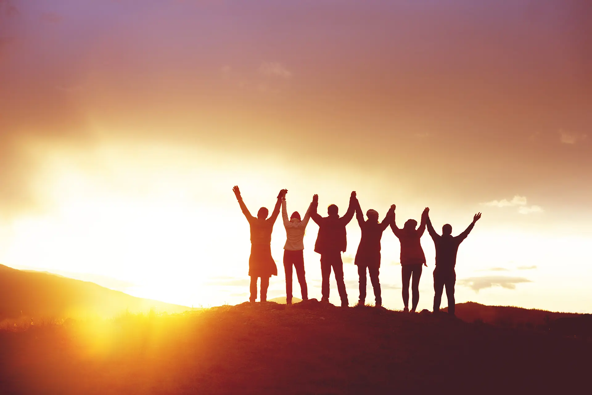 Six people silhouetted against a vivid sunset, holding hands raised in celebration on a hilltop