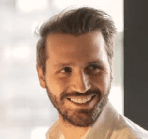 A smiling man with a beard, wearing a white shirt, standing by a window with soft lighting.