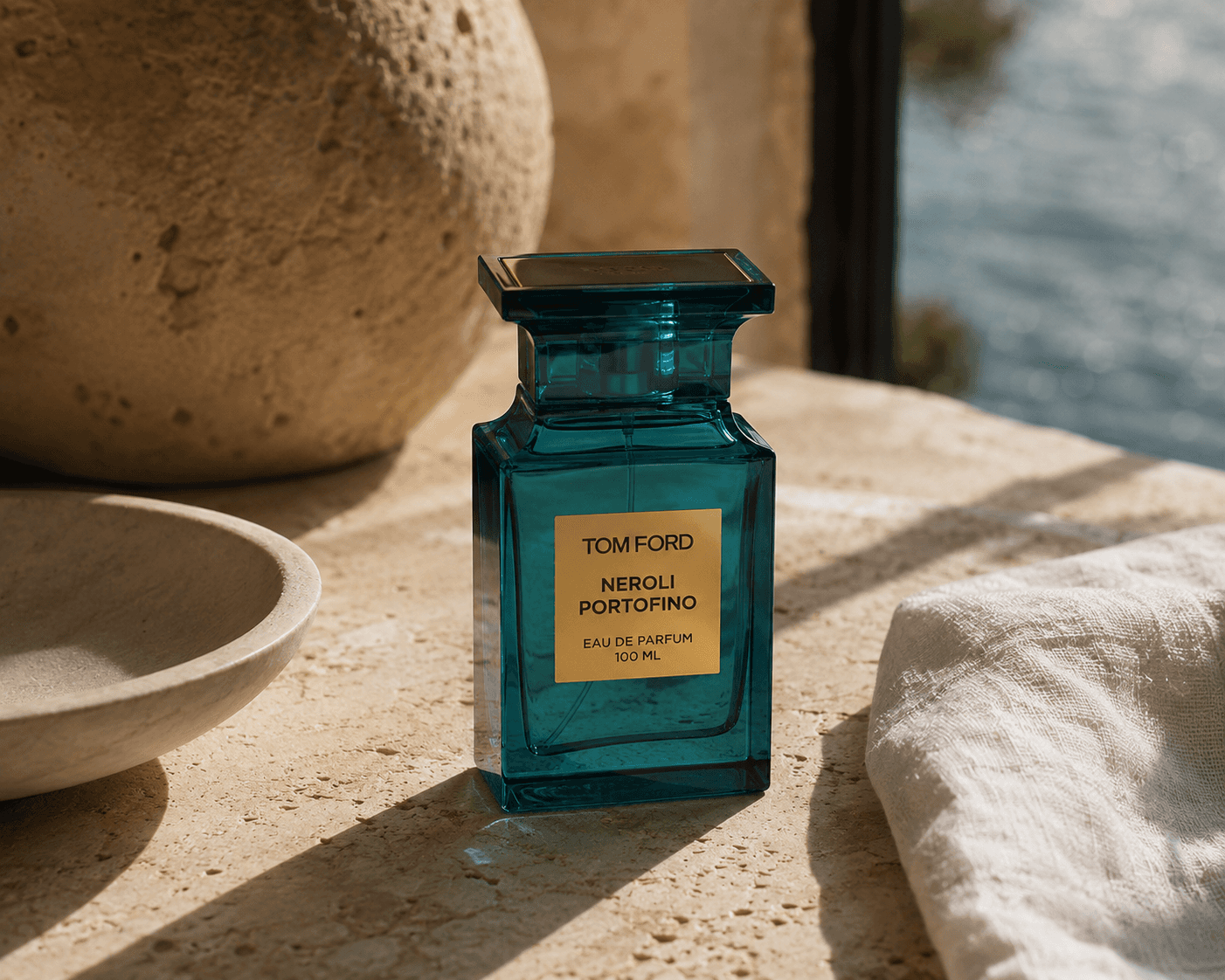 A 100 mL bottle of Tom Ford Neroli Portofino Eau de Parfum in translucent turquoise glass with a gold metallic plaque. The perfume sits on a sunlit beige travertine ledge alongside a ceramic bowl, a round textured stone orb, and draped white linen, with blue Mediterranean water visible through a window in the soft-focus background.