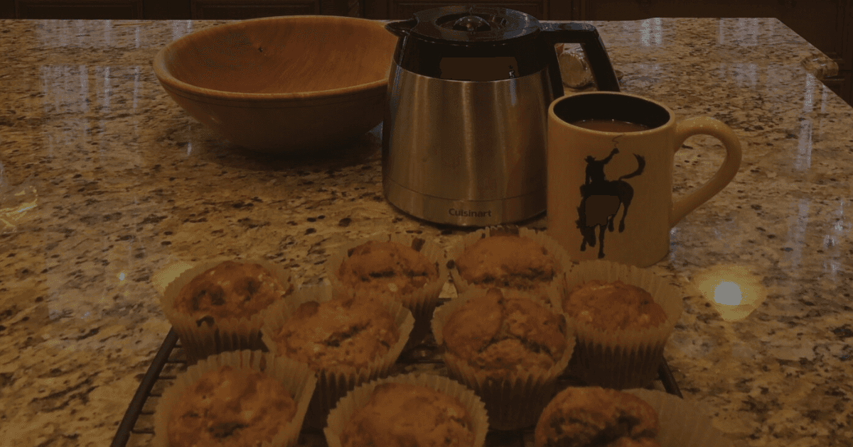 Image of freshly baked muffins on a granite countertop with a carafe of coffee and mug with a bucking bronco etched on the side.
