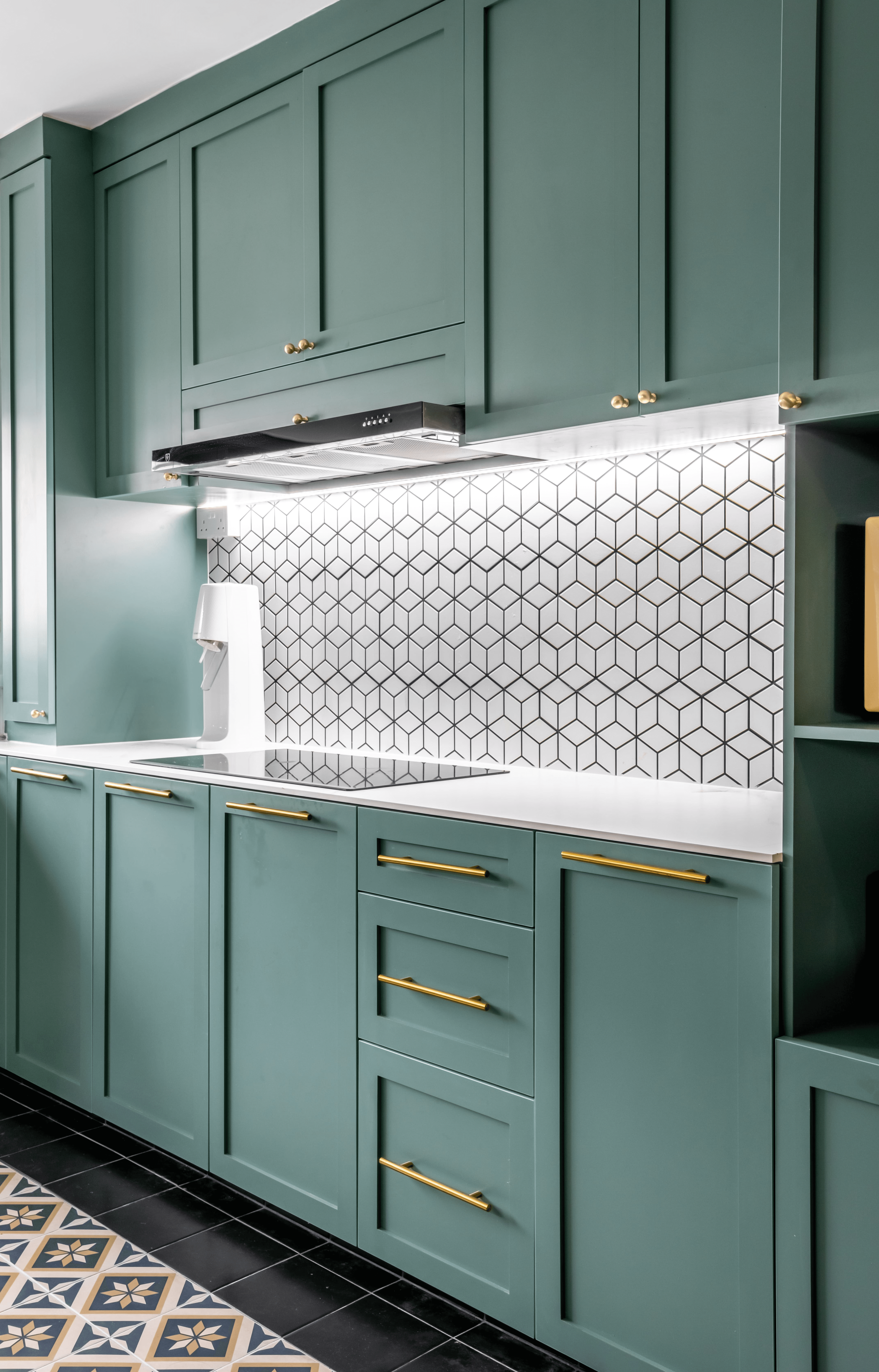 A modern kitchen with matte green cabinets, brass handles, and a geometric black-and-white tiled backsplash under bright lighting.