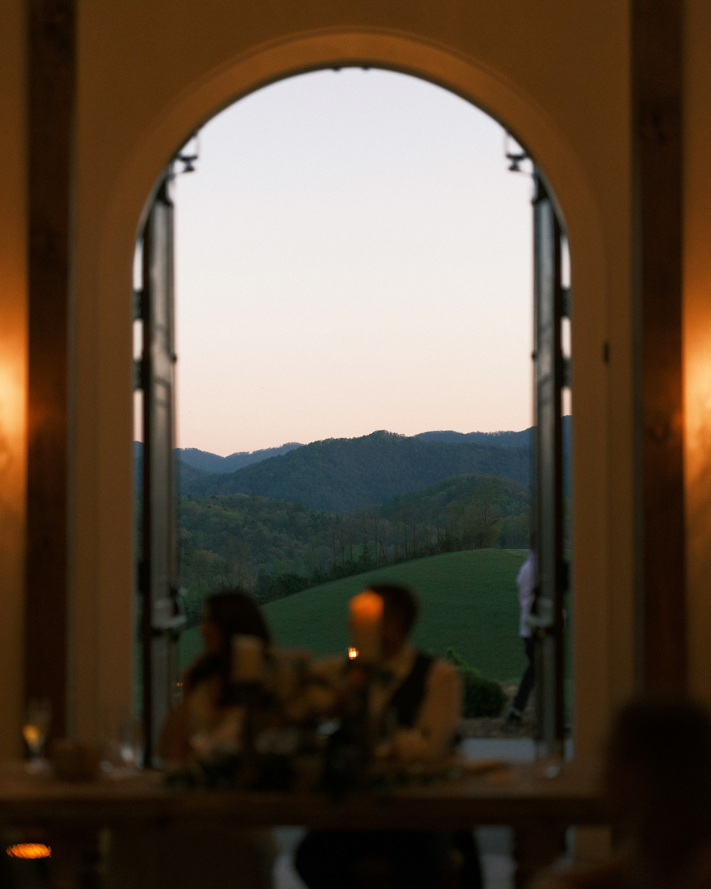 A romantic, softly lit dining setting with blurred foreground figures, showcasing an open arched doorway leading to a serene view of rolling green hills and a pink-hued sunset sky in the background.