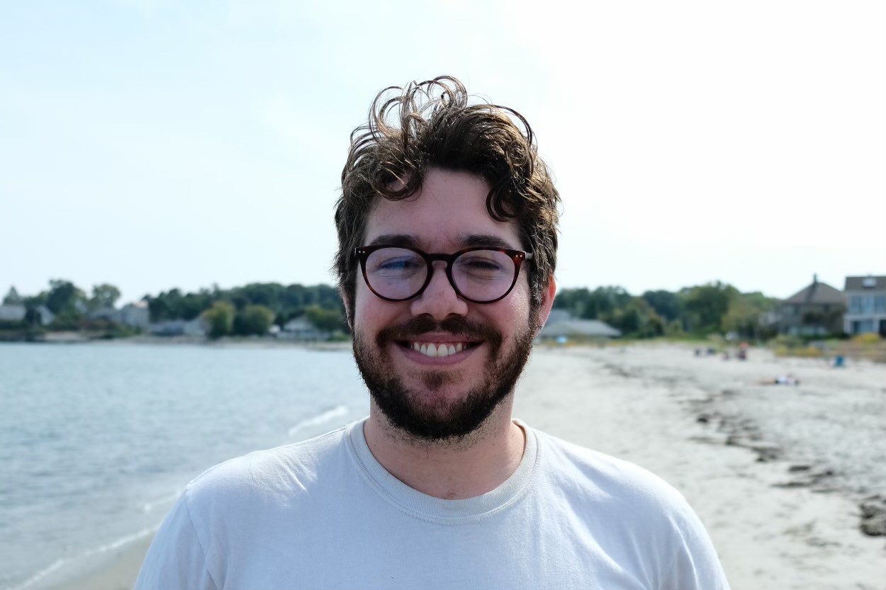 A white man with curly brown hair, a beard, and round glasses, smiling on a beach